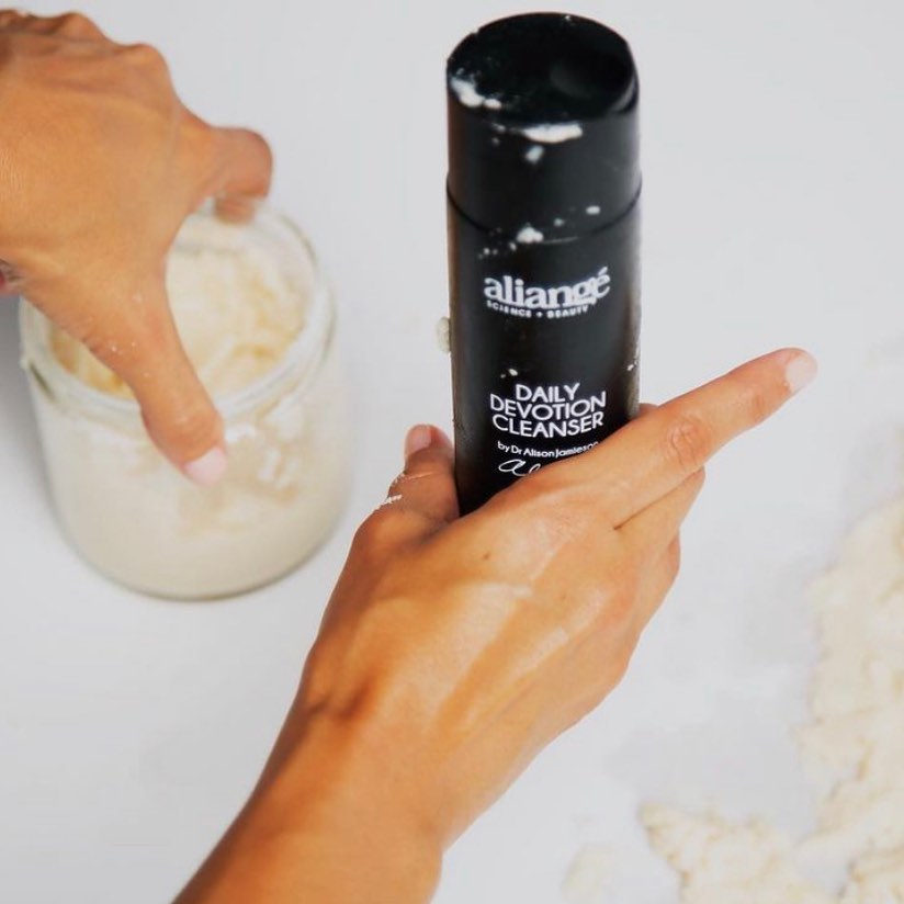 Hands holding the Aliangé Daily Devotion Cleanser above a jar of pale cream — active skincare science