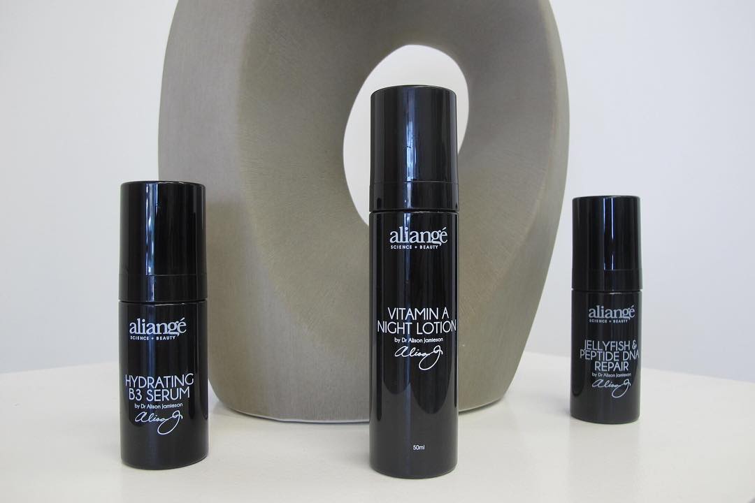 Three Aliangé black bottles grouped beside a pale sculptural vase — Hydrating B3, Vitamin A Night Lotion and Jellyfish Peptide