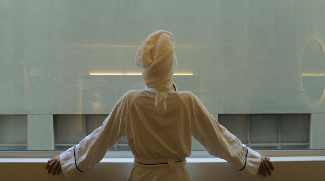Woman in a taupe robe seen from behind with arms outstretched in a bright interior — adult acne article