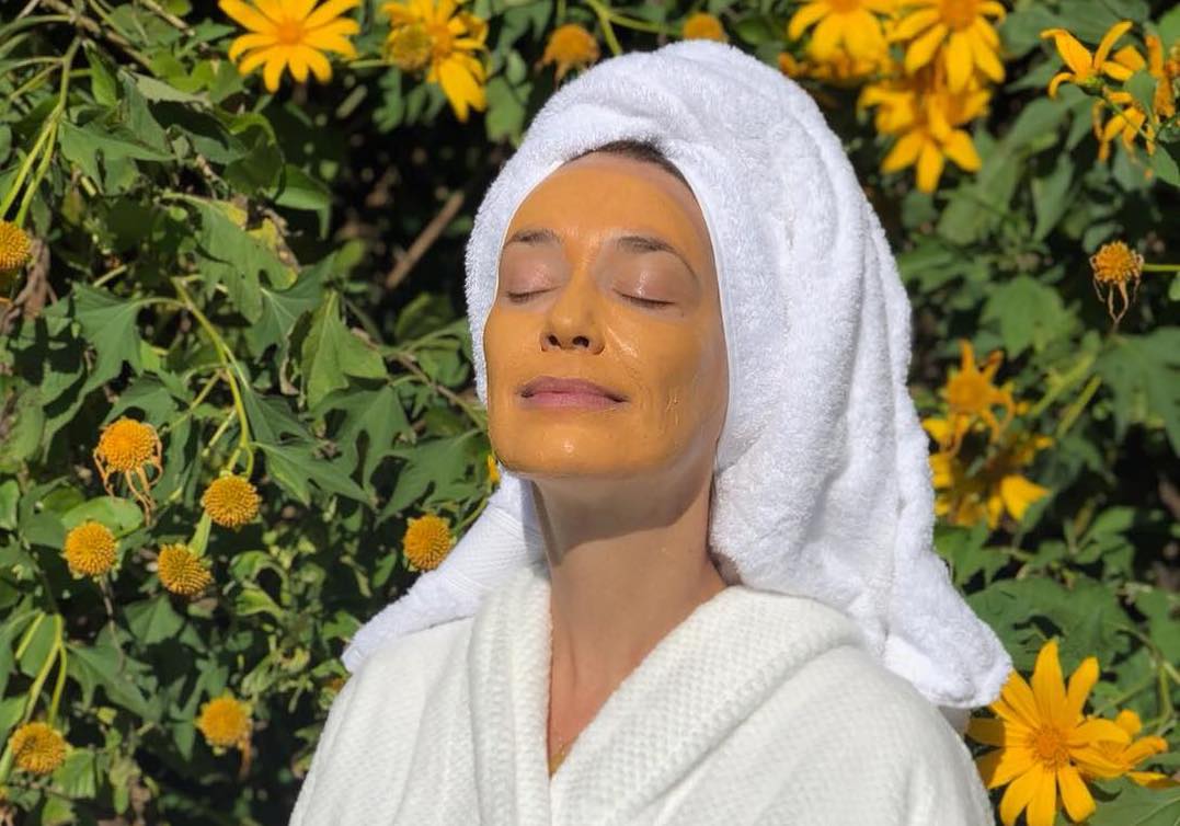 Woman in a white robe and hair towel with a masque on, surrounded by yellow blooms — face masks article